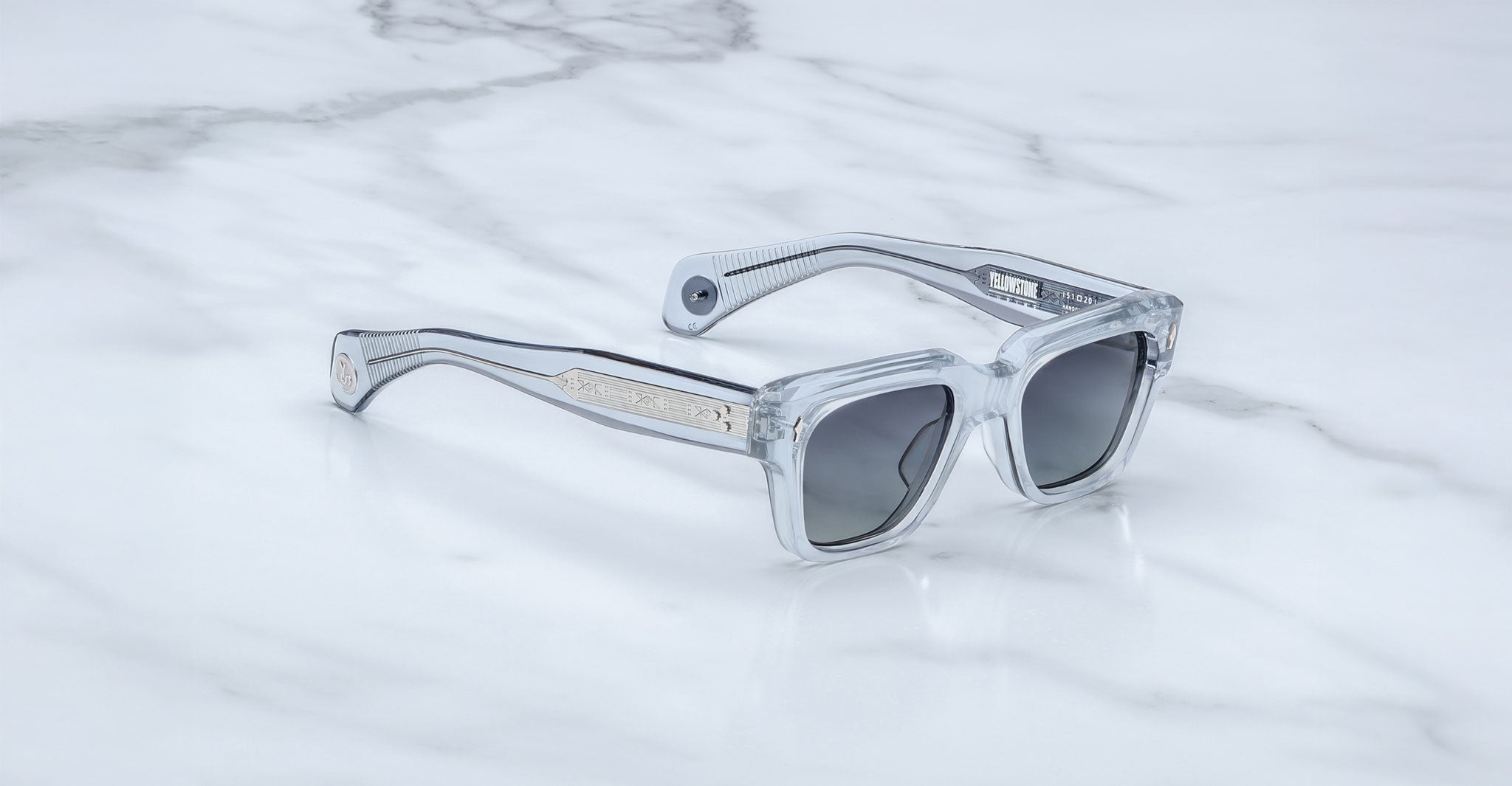 The Jacques Marie Mage Lupine sunglasses, featuring clear frames and dark lenses, rest on a white marble surface. Silver detailing and subtle branding highlight the arms near the hinges.