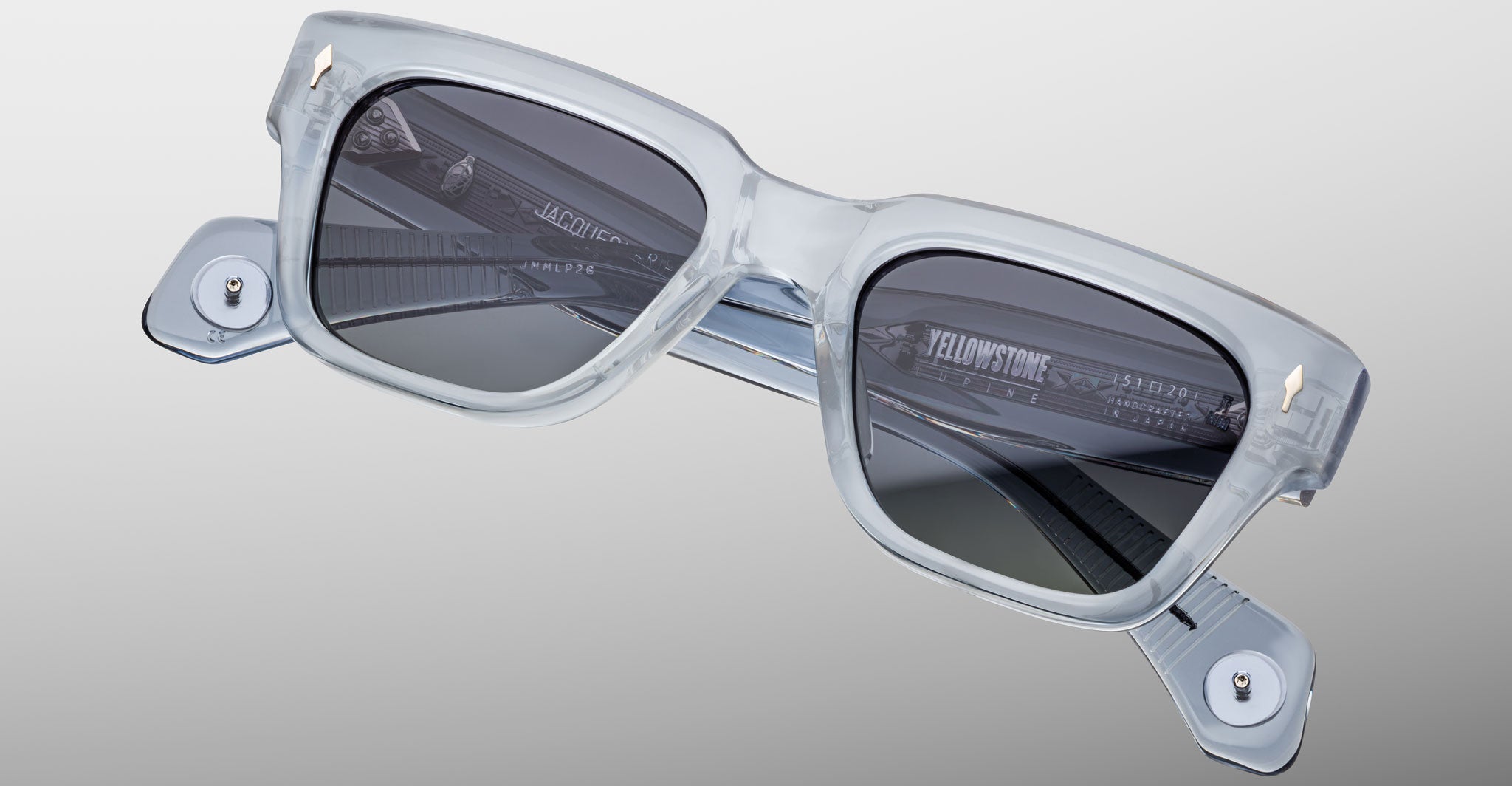 The Jacques Marie Mage Lupine sunglasses feature square, thick transparent light gray frames, dark lenses, metallic hinge accents, and visible interior branding, all set against a simple light background.