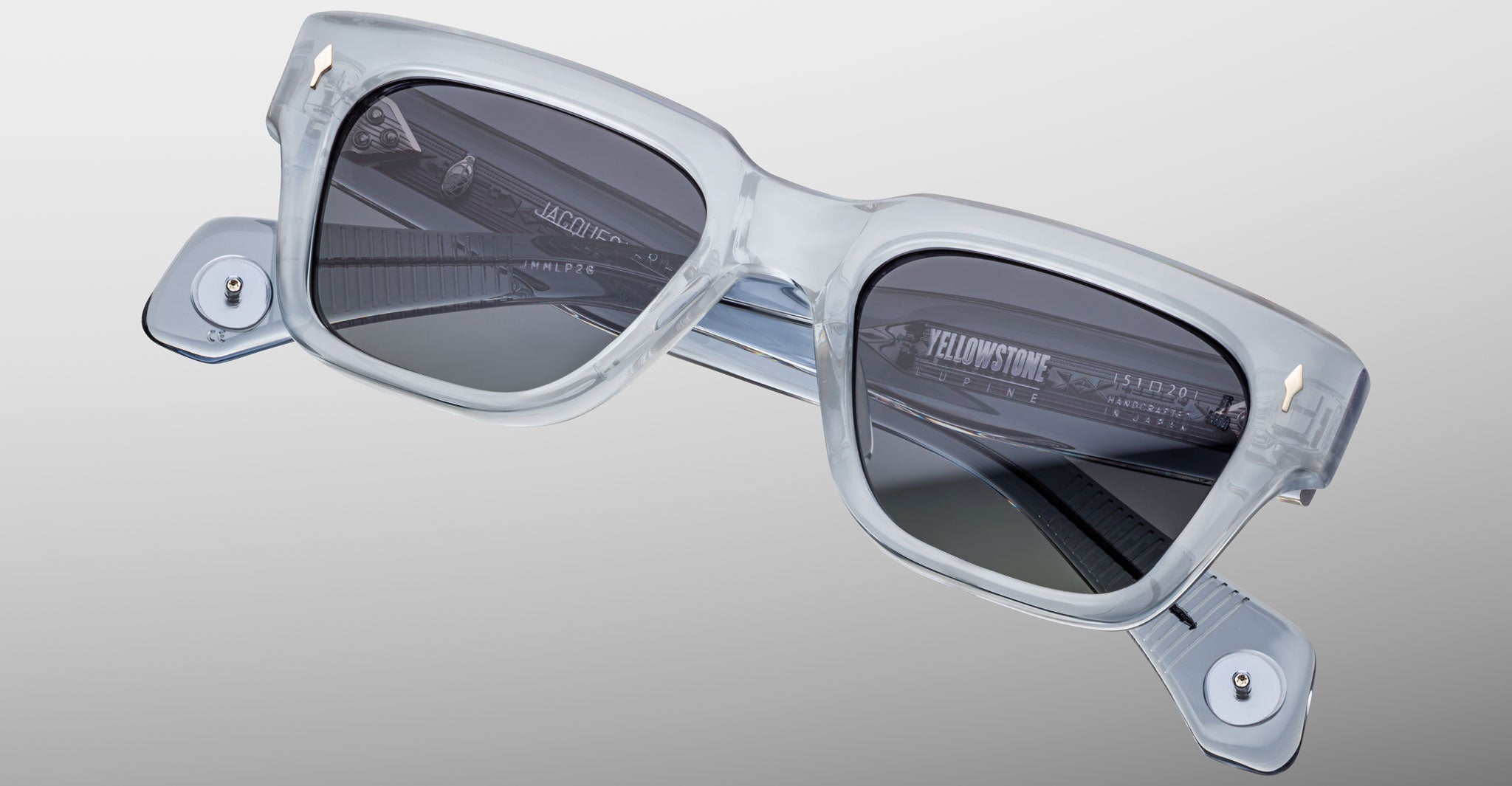 The Jacques Marie Mage Lupine sunglasses feature square, thick transparent light gray frames, dark lenses, metallic hinge accents, and visible interior branding, all set against a simple light background.
