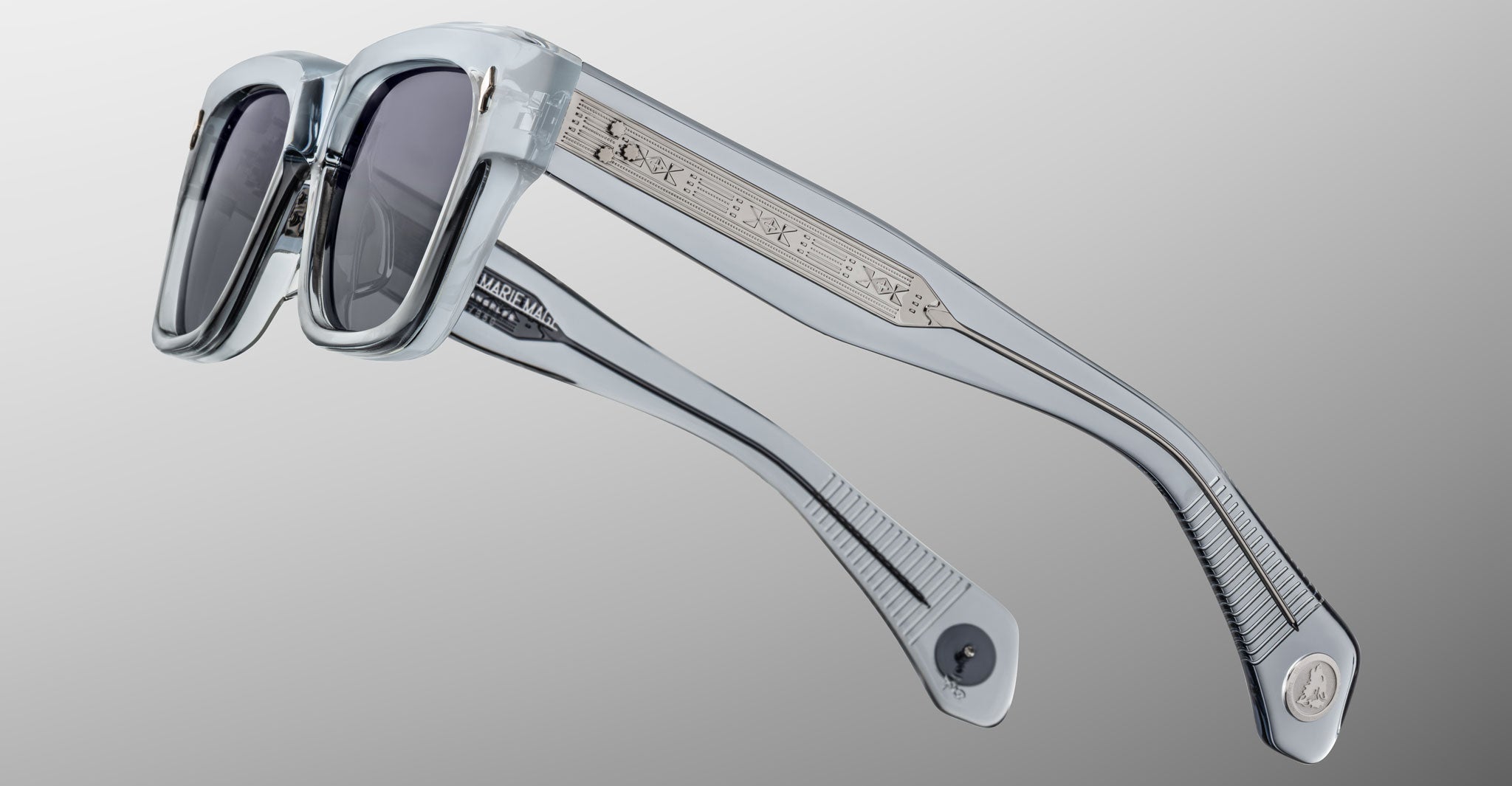 The Jacques Marie Mage Lupine sunglasses, shown at an angle, feature clear square frames with dark lenses, metallic accents on the temples, and engraved details, set against a smooth grey gradient background.