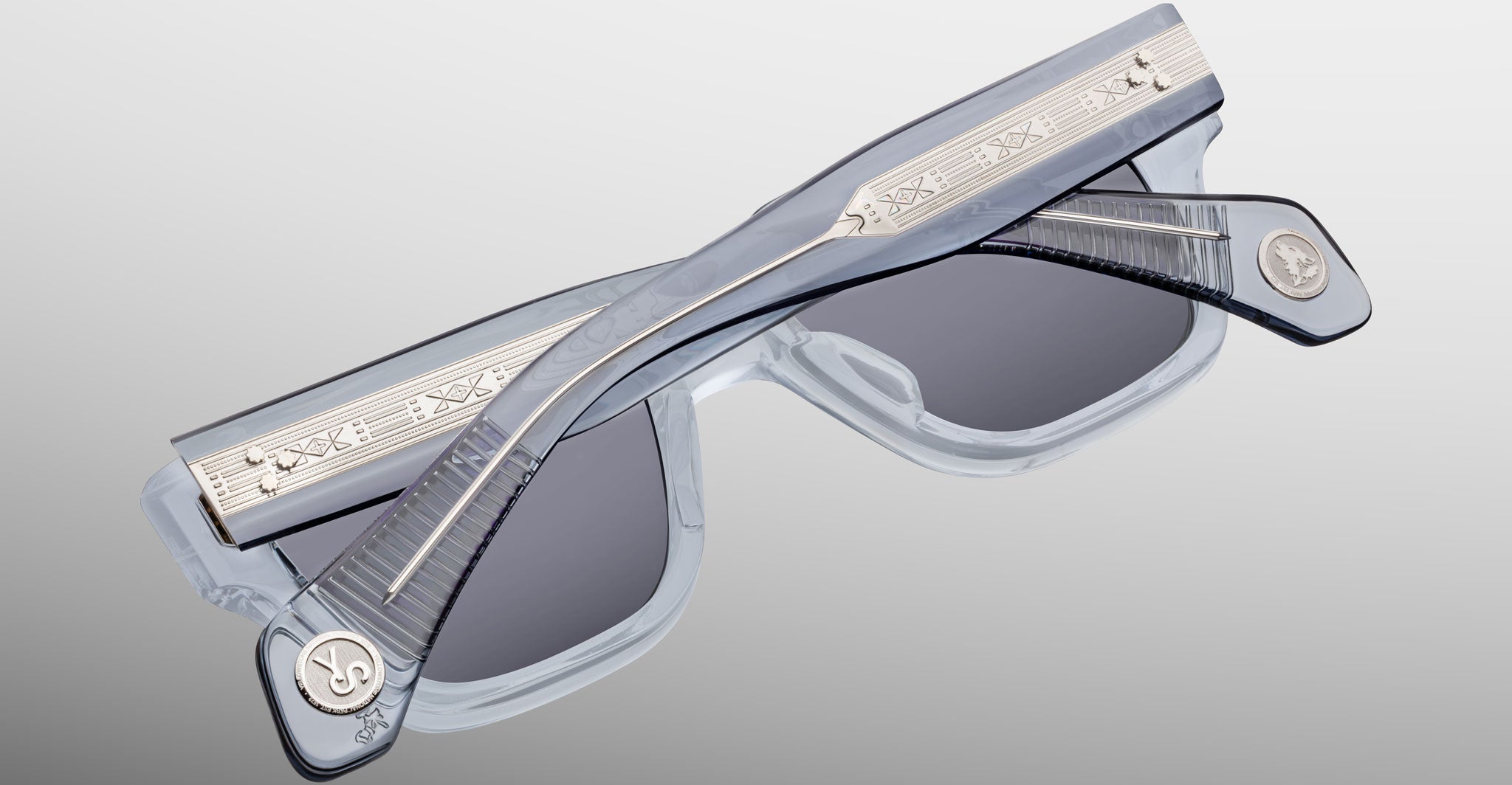 Jacques Marie Mage Lupine sunglasses, shown folded, feature clear frames with dark lenses. The temples include silver-tone metal detailing with engraved patterns and round emblems near the hinges.