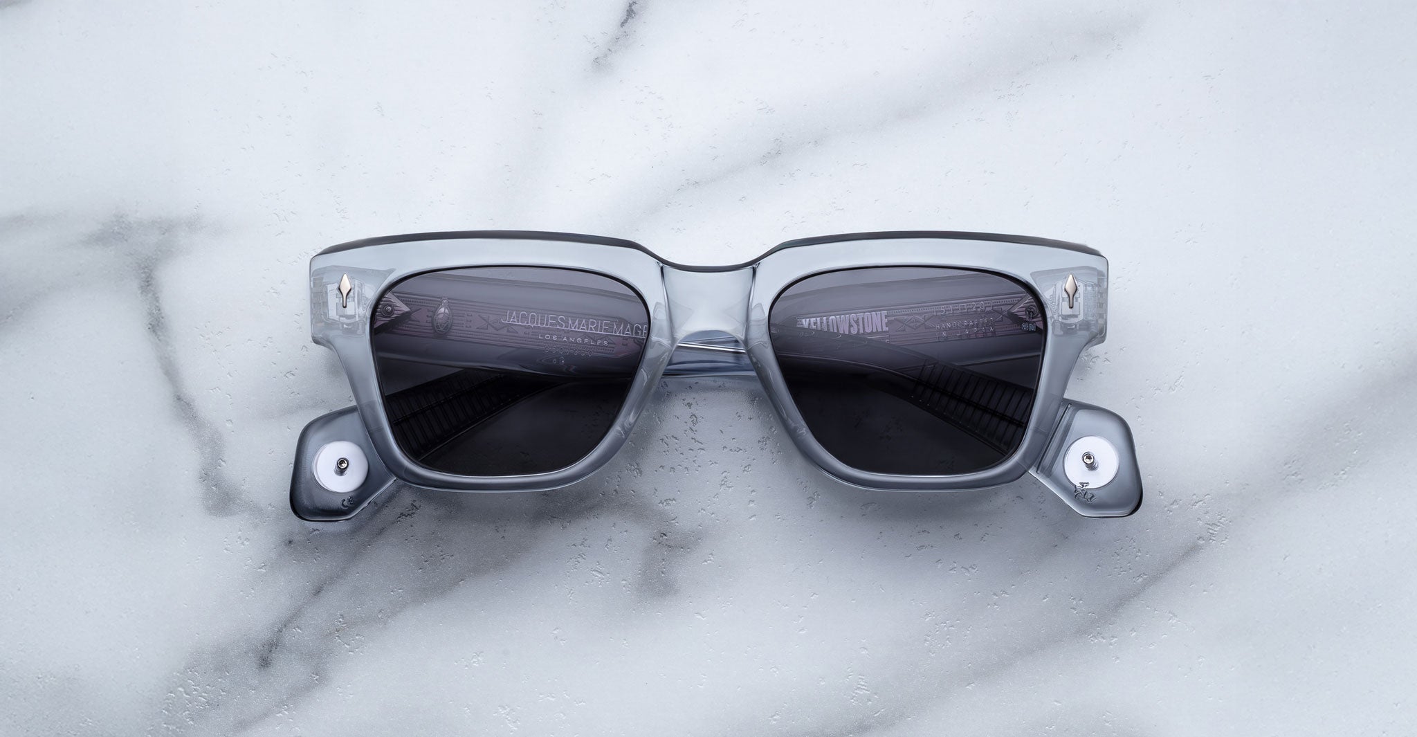 A pair of Jacques Marie Mage Lupine rectangular gray transparent sunglasses with dark lenses rests on a white marble surface.