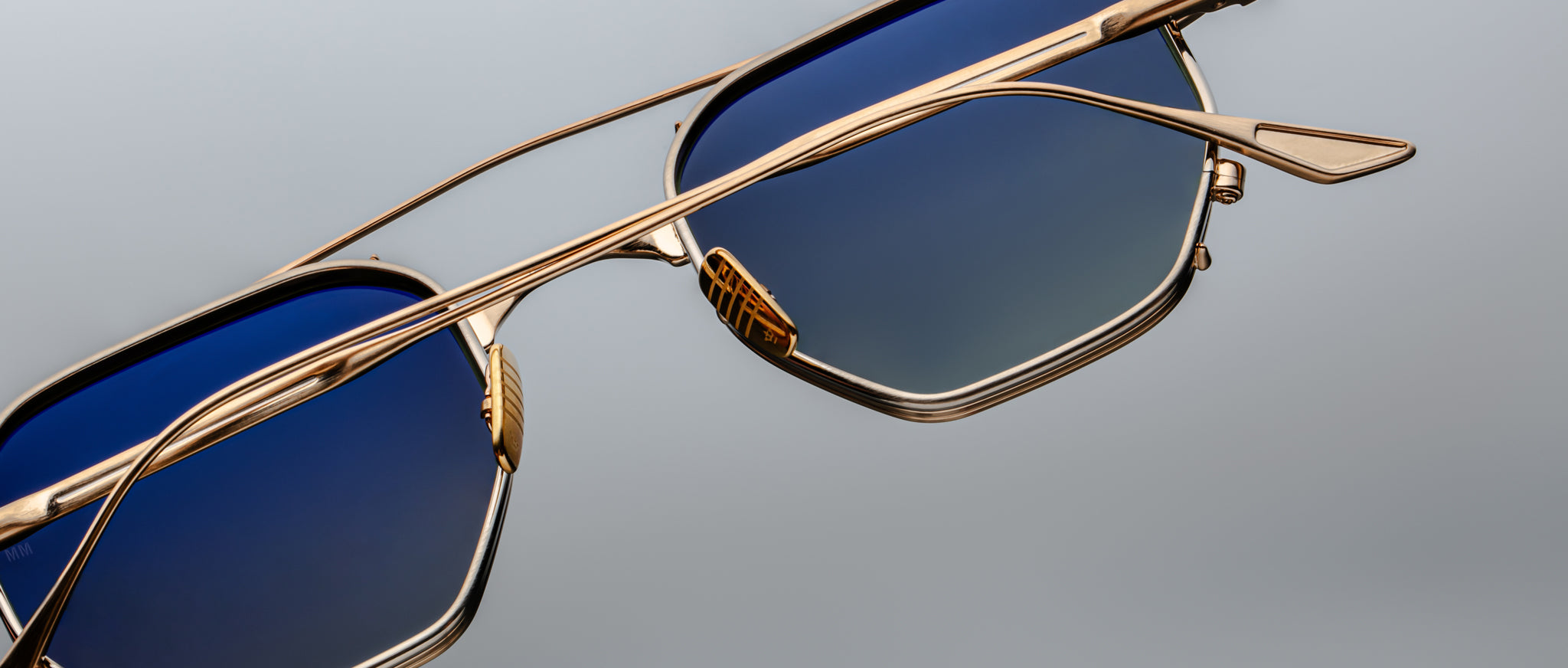 Close-up of Jacques Marie Mage Marbot sunglasses featuring gold metal frames and blue gradient lenses, set against a plain gray background.