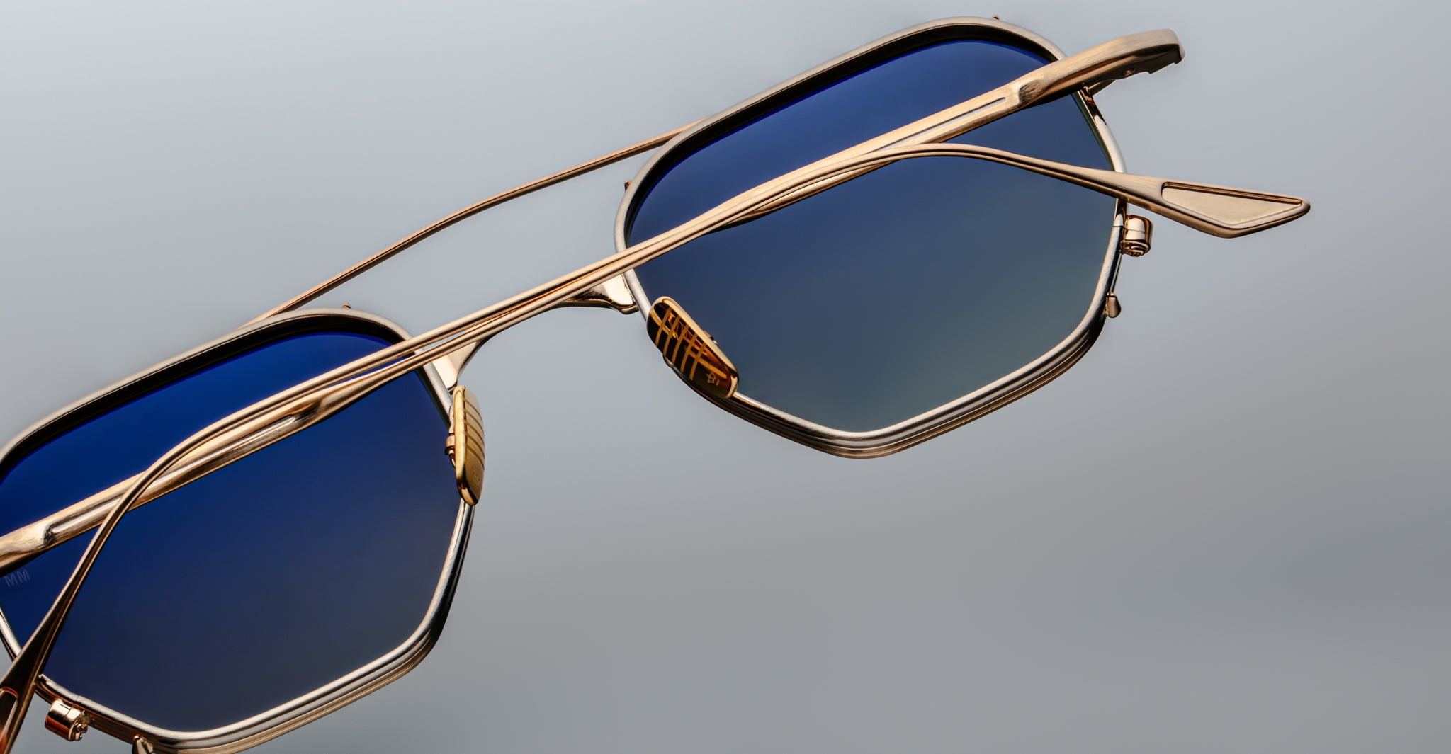 Close-up of Jacques Marie Mage Marbot sunglasses featuring gold metal frames and blue gradient lenses, set against a plain gray background.