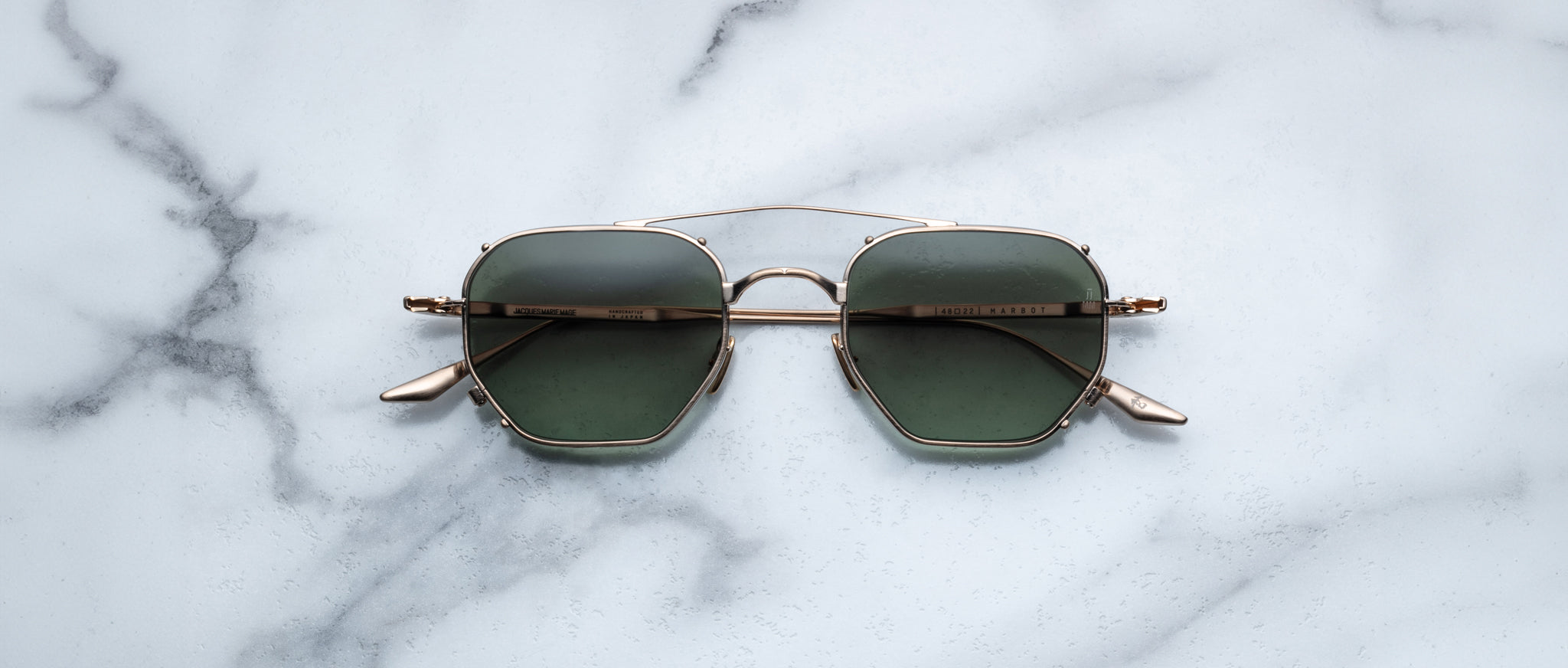 A pair of Jacques Marie Mage Marbot gold-framed sunglasses with green lenses rests on a white marble surface streaked with gray veining.