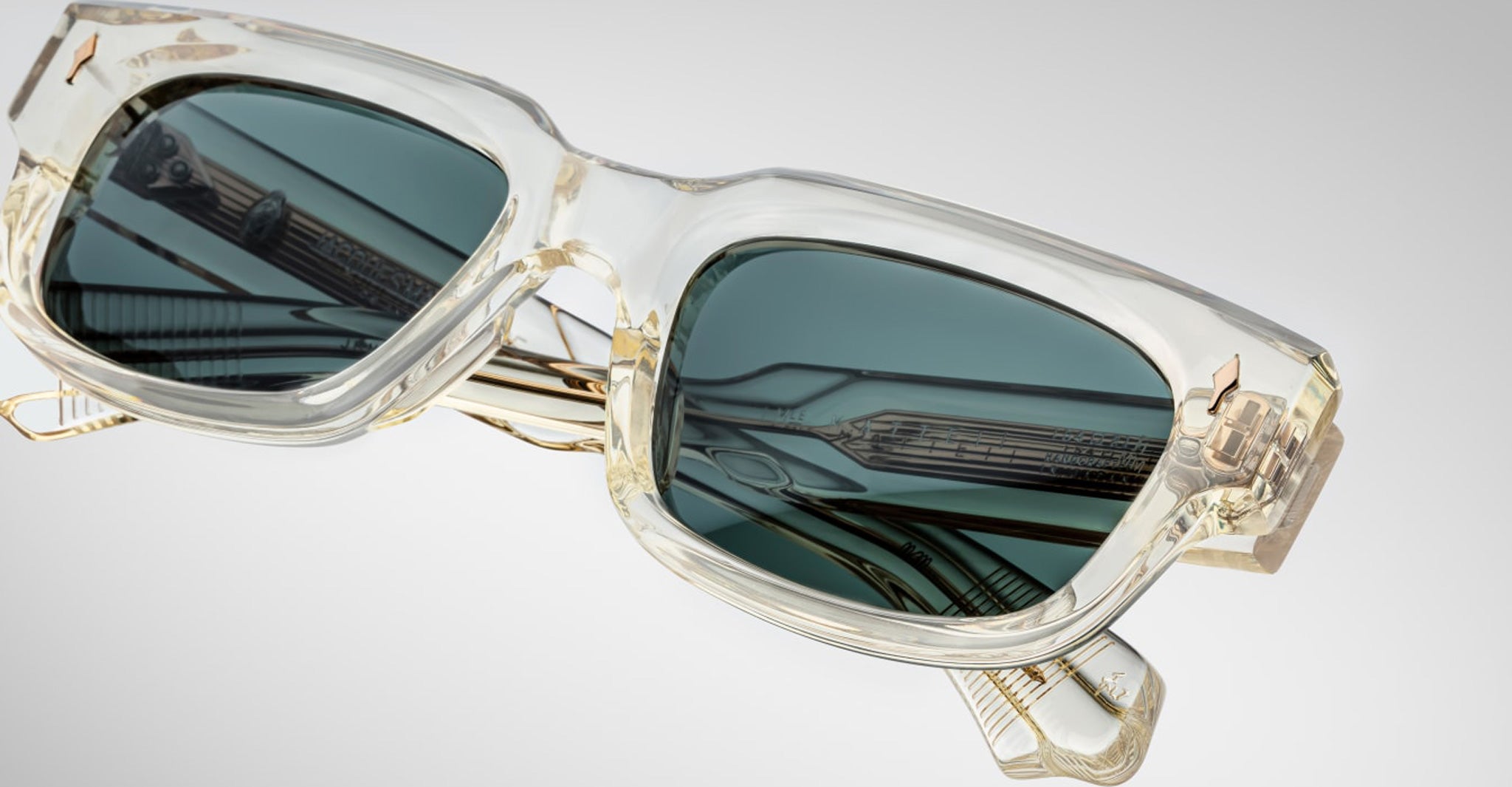 Close-up of Jacques Marie Mage Mattei sunglasses: stylish, rectangular design with clear, thick frames, dark tinted lenses, and temples featuring metallic details and intricate patterns against a light gray background.