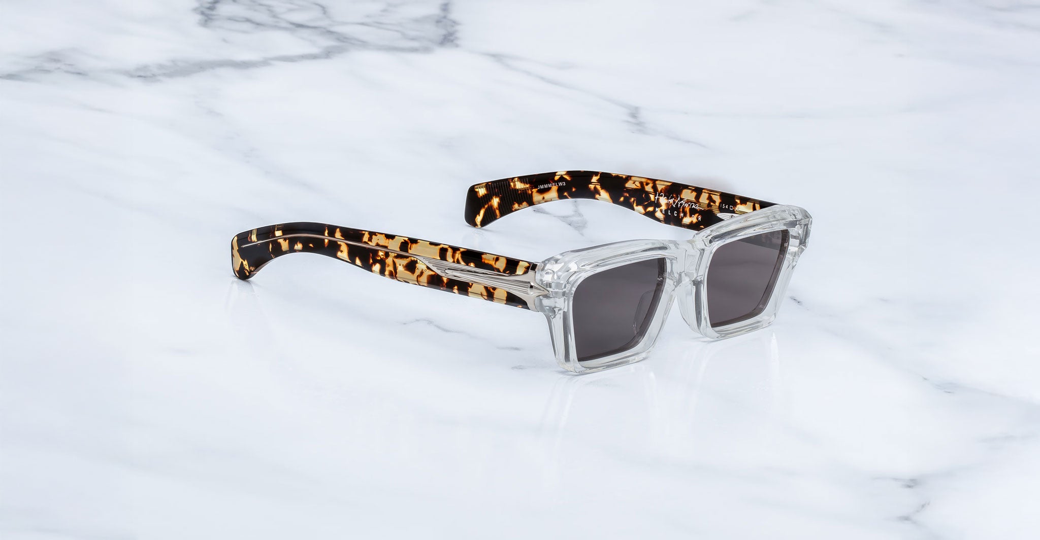Jacques Marie Mage Melchior sunglasses with clear frames, UV-protected dark lenses, and tortoiseshell arms, front view.