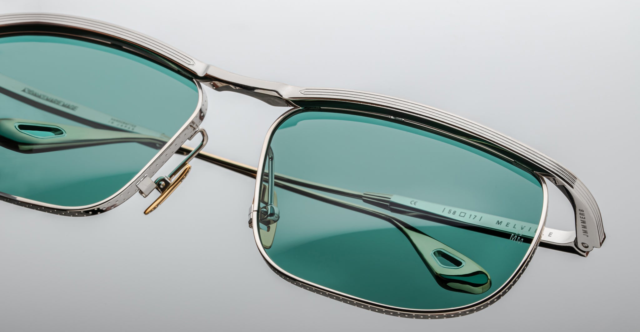 Close-up of Jacques Marie Mage Melville eyeglasses featuring green-tinted lenses, a silver metal frame, and intricate detailing on the brow bar and temples, catching light on a smooth, pale surface.