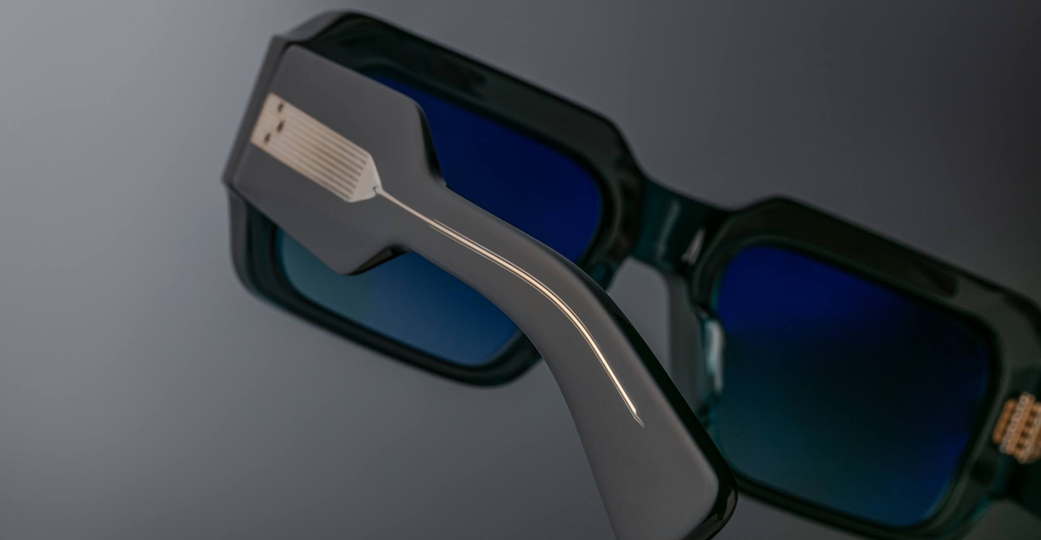 Close-up of Jacques Marie Mage Miglia sunglasses in black with blue lenses, highlighting the thick frame and intricate temple pattern, set against a gradient gray background.