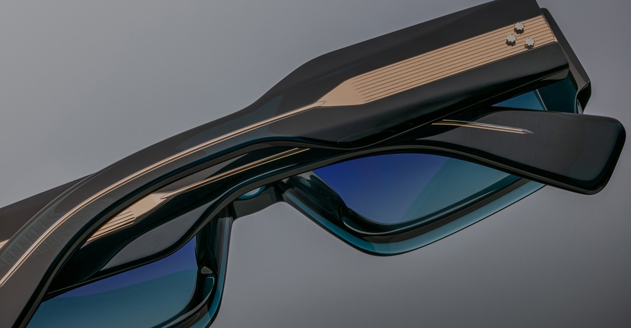A close-up of two pairs of Jacques Marie Mage Miglia sunglasses with dark frames and blue-tinted lenses, stacked together, highlighting their metallic accents and detailed arms.
