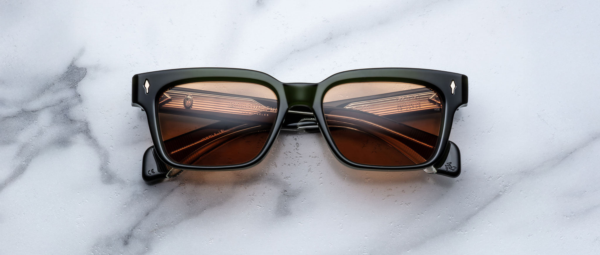 A pair of Jacques Marie Mage Molino 55 black square-framed sunglasses with brown lenses rests on a white and gray marble surface, the lenses reflecting lights and a ceiling structure.