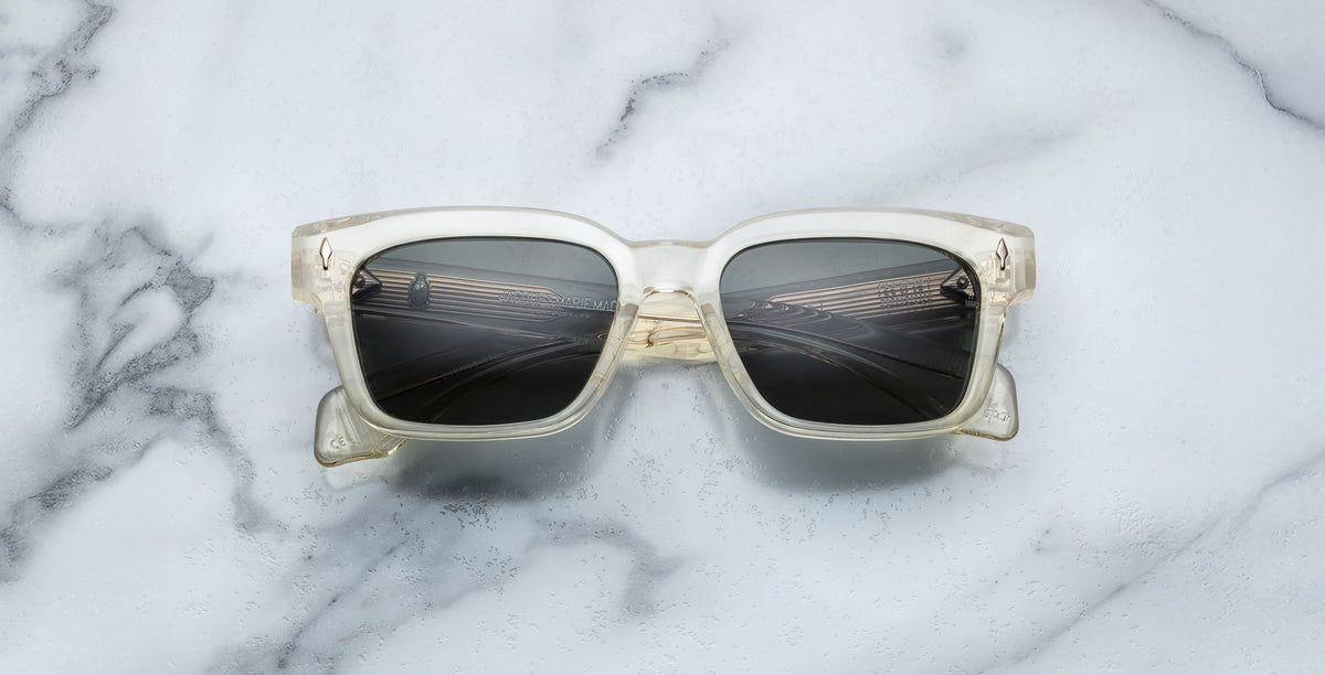 The Jacques Marie Mage Molino 55 sunglasses with clear square frames and dark lenses are centered on a white marble surface with gray veins, casting a faint shadow.