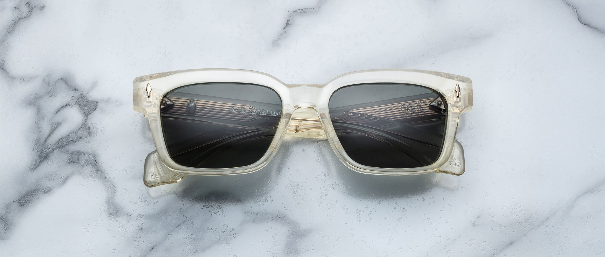The Jacques Marie Mage Molino 55 sunglasses with clear square frames and dark lenses are centered on a white marble surface with gray veins, casting a faint shadow.