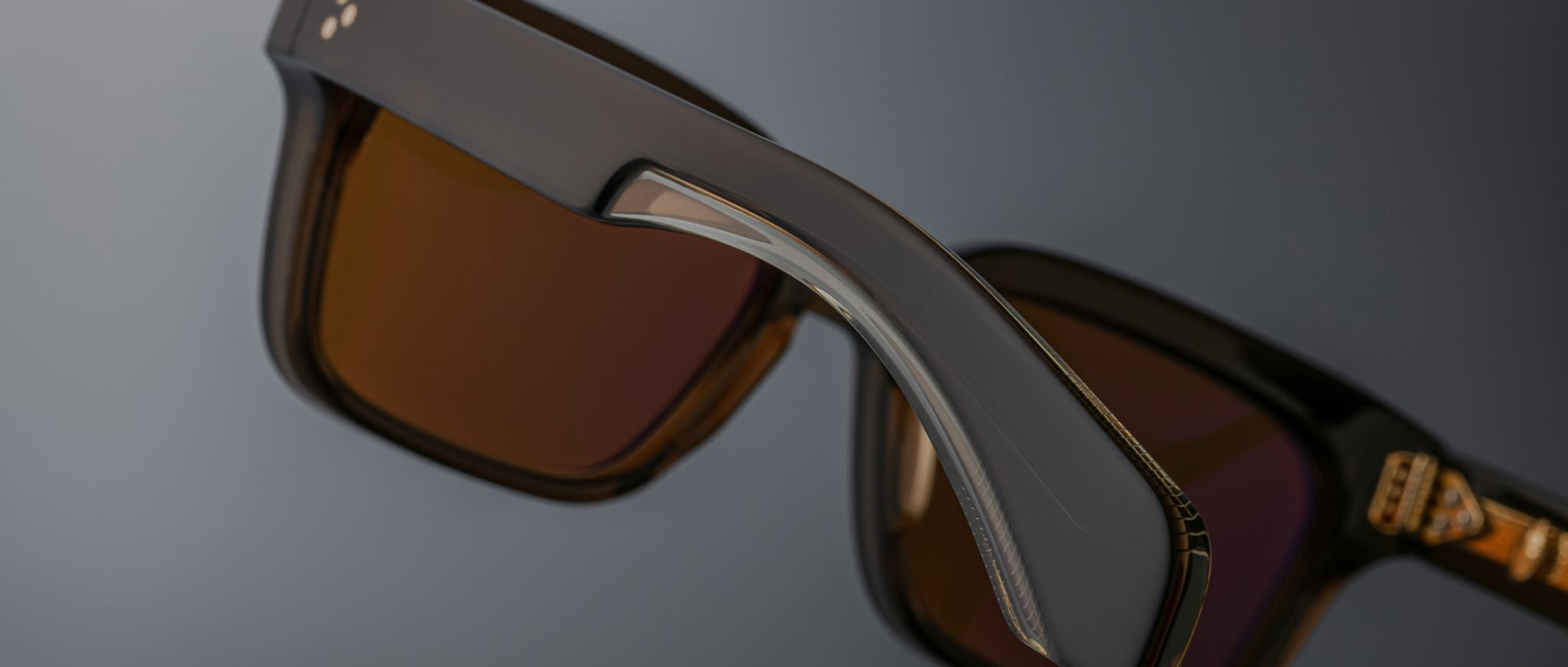 Close-up of Jacques Marie Mage Molino 55 sunglasses featuring thick dark brown frames and amber-tinted lenses, with focus on the arm and hinge, set against a blurred gray background.