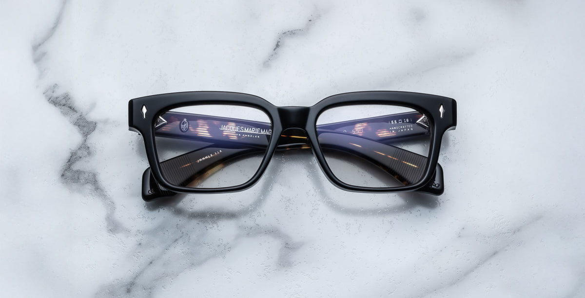 A pair of Jacques Marie Mage Molino 55 black rectangular eyeglasses rests on a white marble surface, with digital tunnel and interface graphics reflected on the lenses.