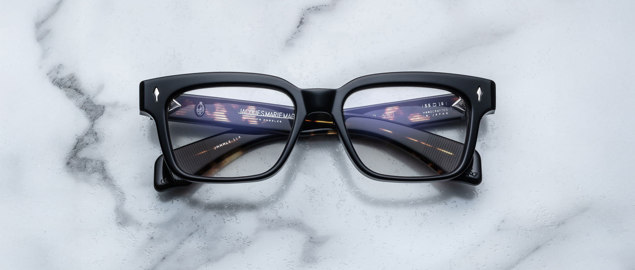 A pair of Jacques Marie Mage Molino 55 black rectangular eyeglasses rests on a white marble surface, with digital tunnel and interface graphics reflected on the lenses.