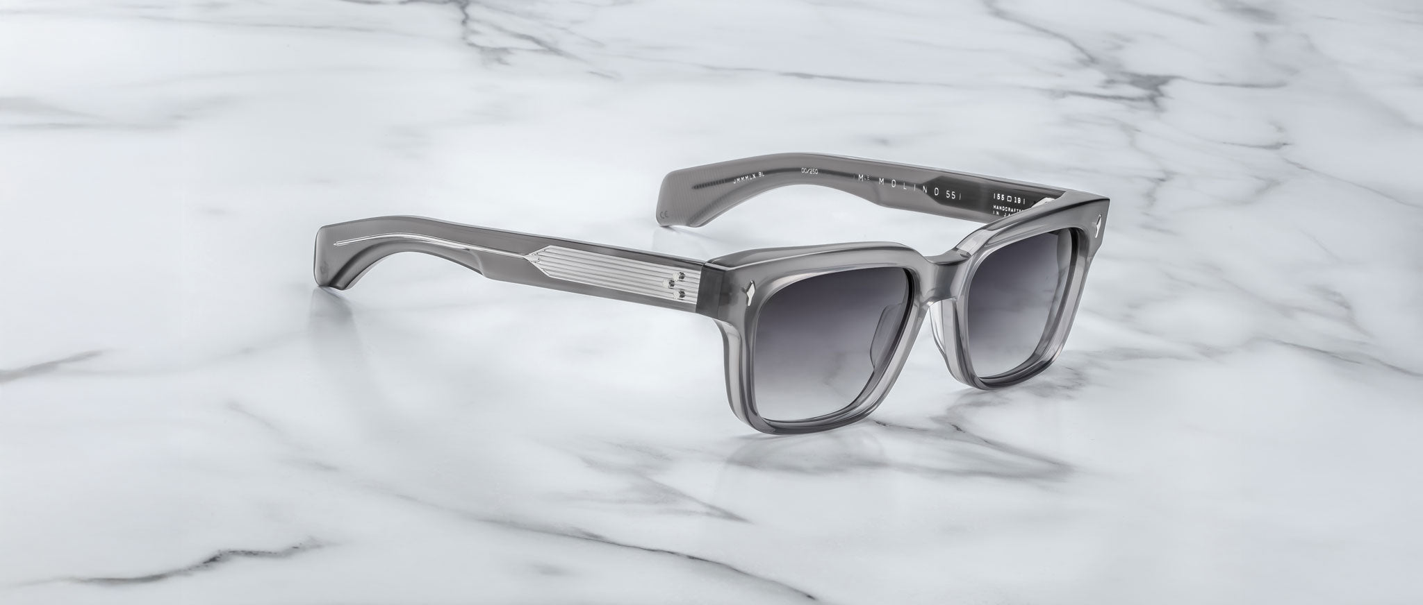 A pair of Jacques Marie Mage Molino 55 gray square-frame sunglasses with dark lenses rests on a white marble surface.