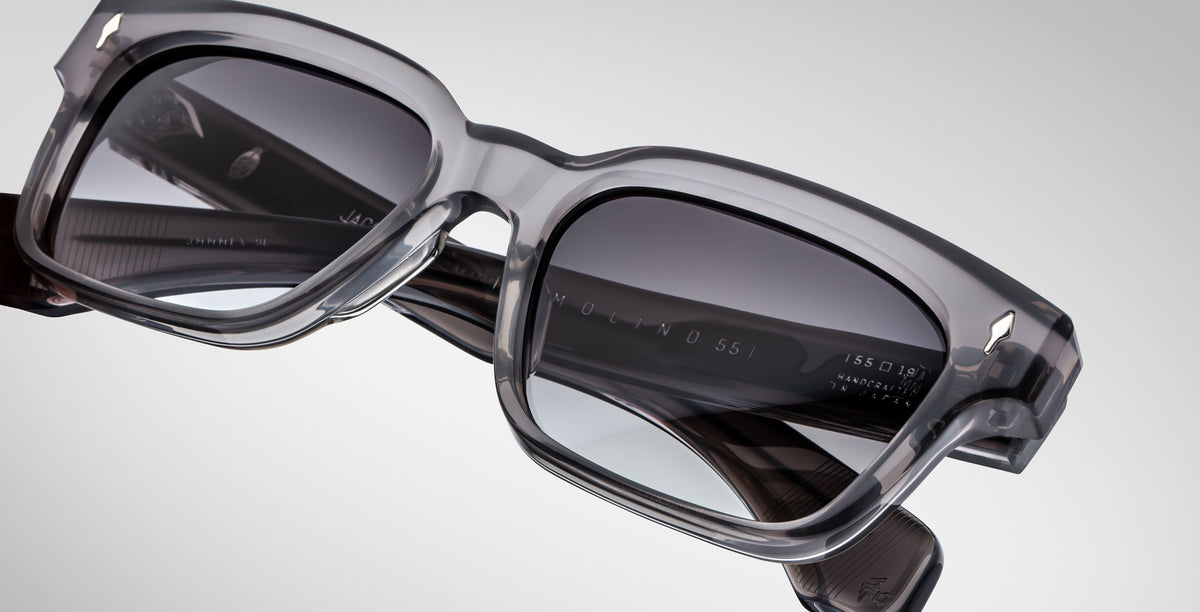 Close-up of Jacques Marie Mage Molino 55 sunglasses—stylish, rectangular gray eyewear with thick, transparent frames and dark-tinted lenses set against a plain, light background.