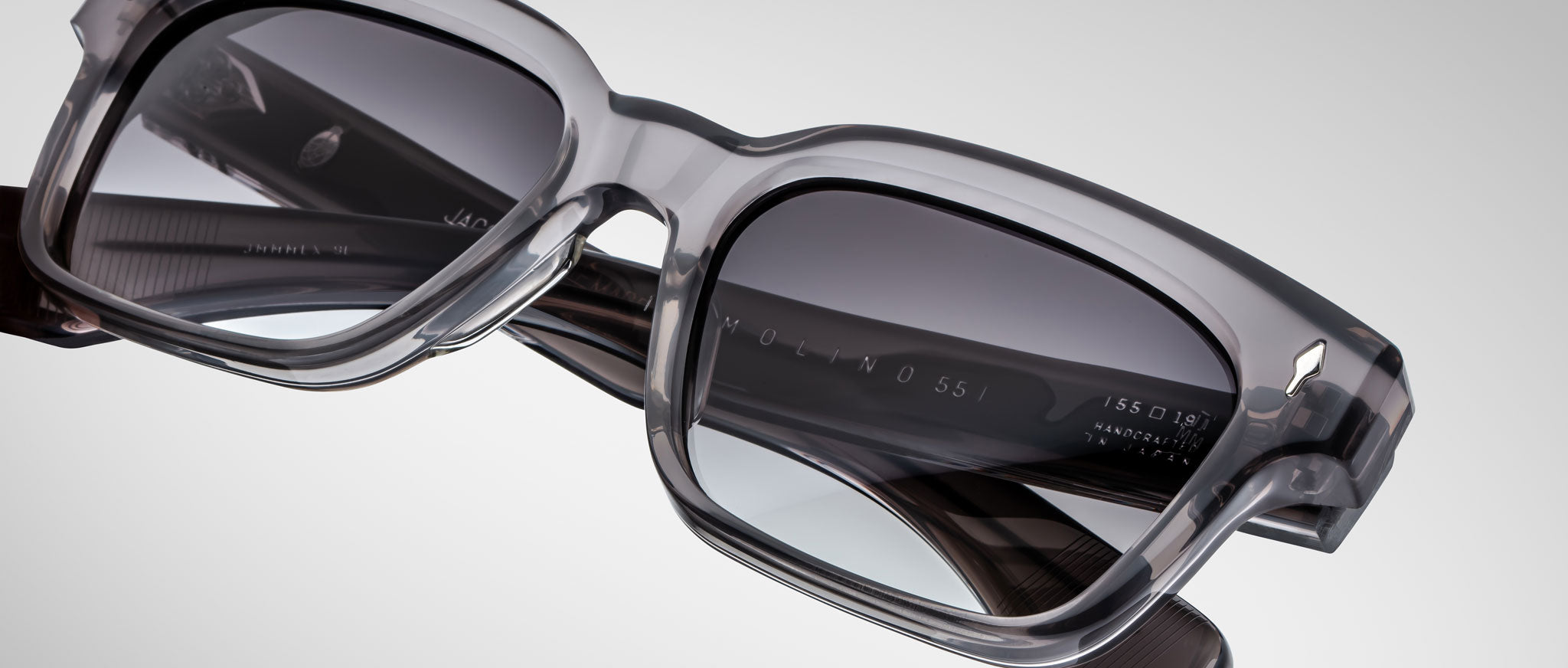 Close-up of Jacques Marie Mage Molino 55 sunglasses—stylish, rectangular gray eyewear with thick, transparent frames and dark-tinted lenses set against a plain, light background.