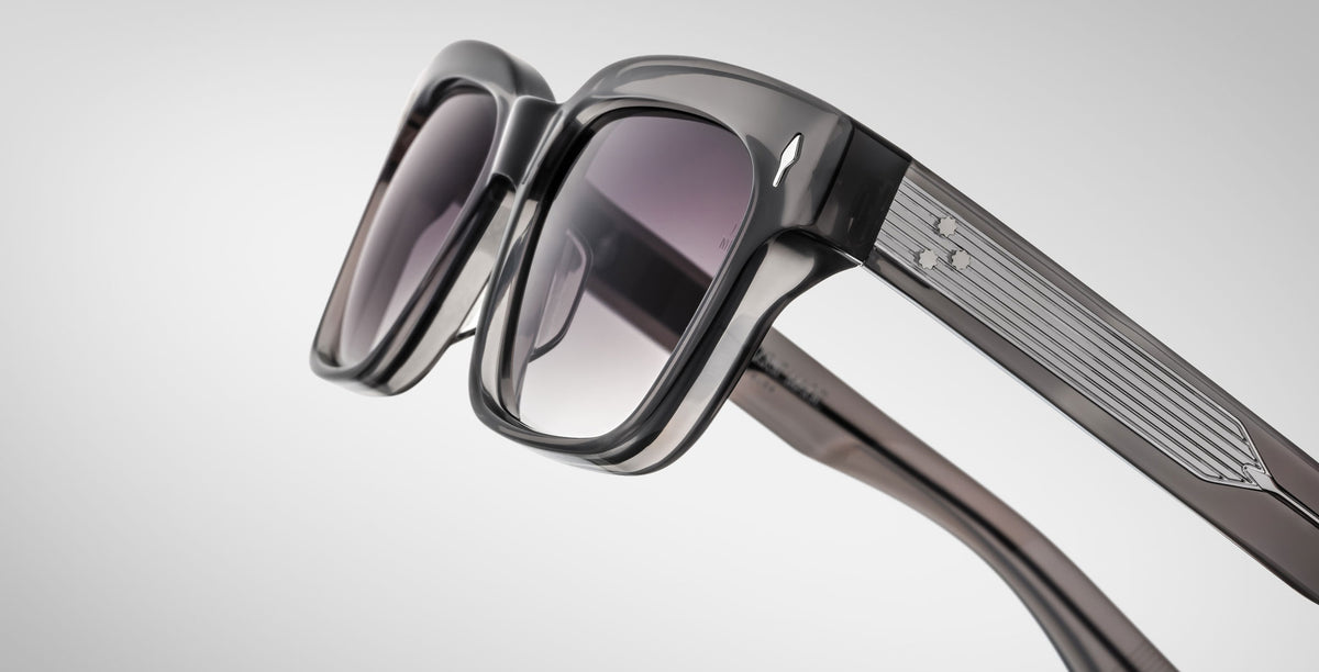 Close-up of Jacques Marie Mage Molino 55 sunglasses featuring stylish thick black frames, slightly tinted lenses, and metallic detailing on the arms against a light gray background.
