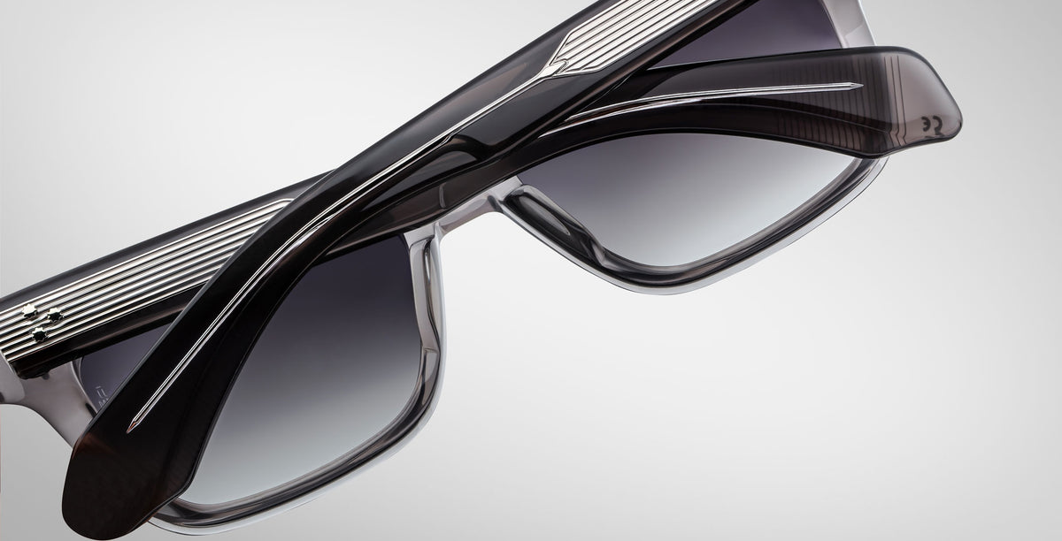 A close-up of two pairs of Jacques Marie Mage Molino 55 sunglasses with thick, dark frames and gradient lenses, overlapping each other against a light gray background.
