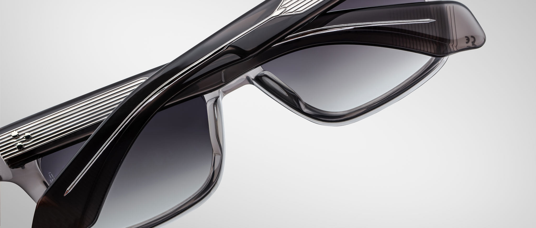 A close-up of two pairs of Jacques Marie Mage Molino 55 sunglasses with thick, dark frames and gradient lenses, overlapping each other against a light gray background.