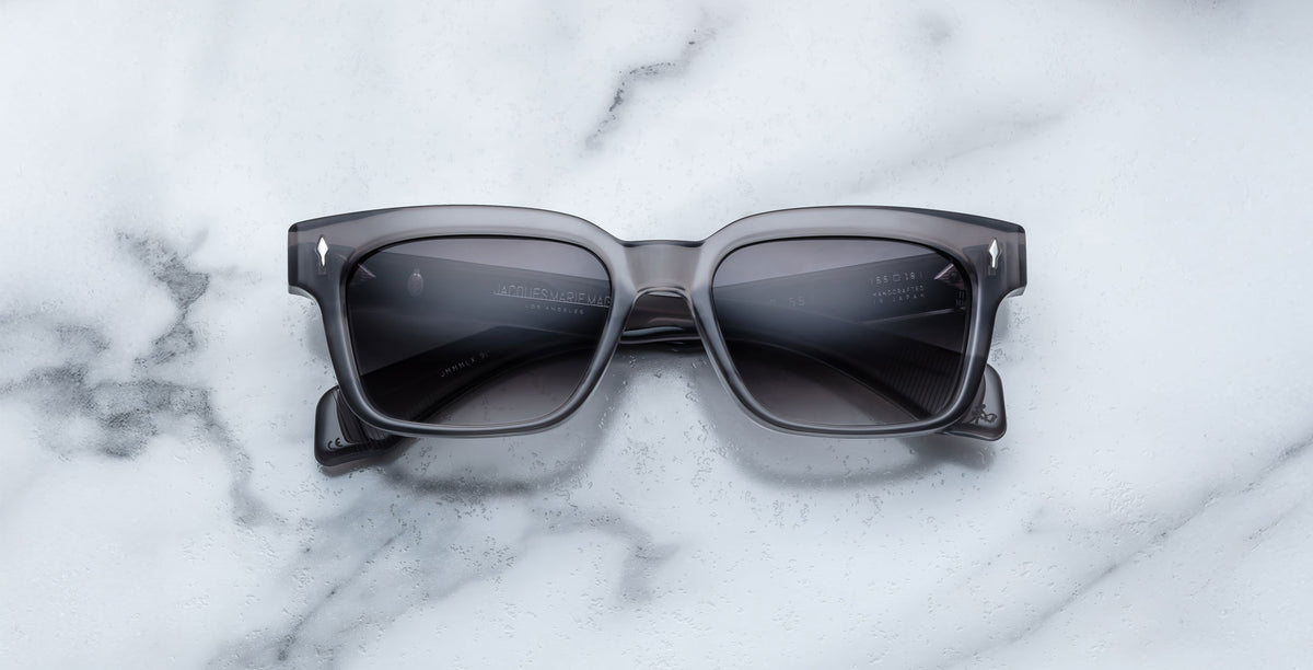 A pair of Jacques Marie Mage Molino 55 black rectangular sunglasses with dark lenses rests on a white marble surface with gray veining.