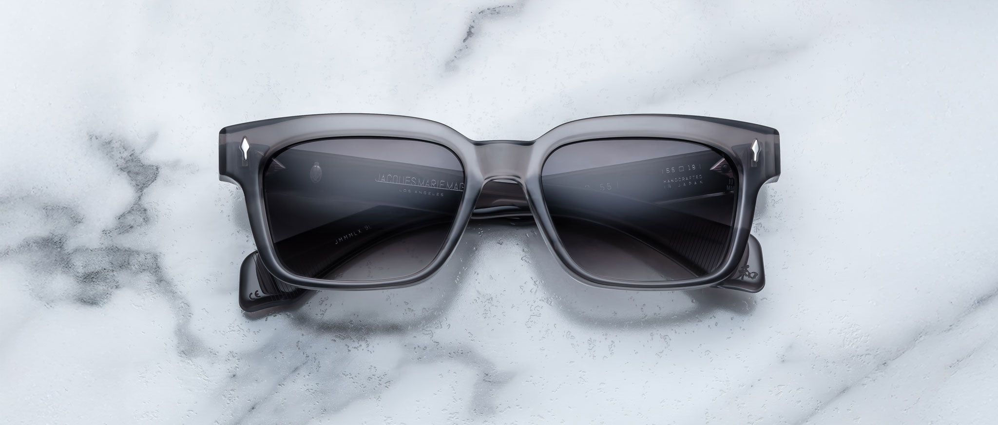 A pair of Jacques Marie Mage Molino 55 black rectangular sunglasses with dark lenses rests on a white marble surface with gray veining.