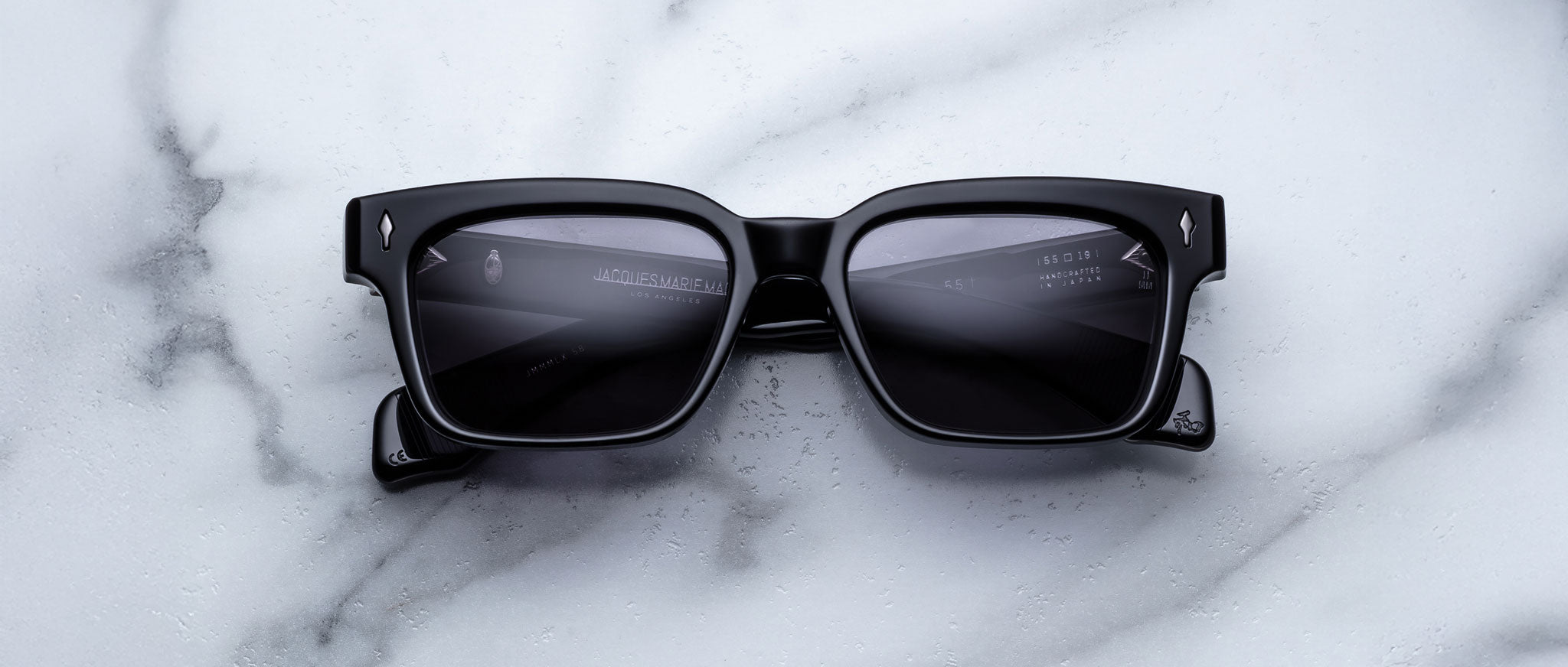 A pair of Jacques Marie Mage Molino 55 black rectangular sunglasses with dark lenses rests on a white marble surface with grey veining.