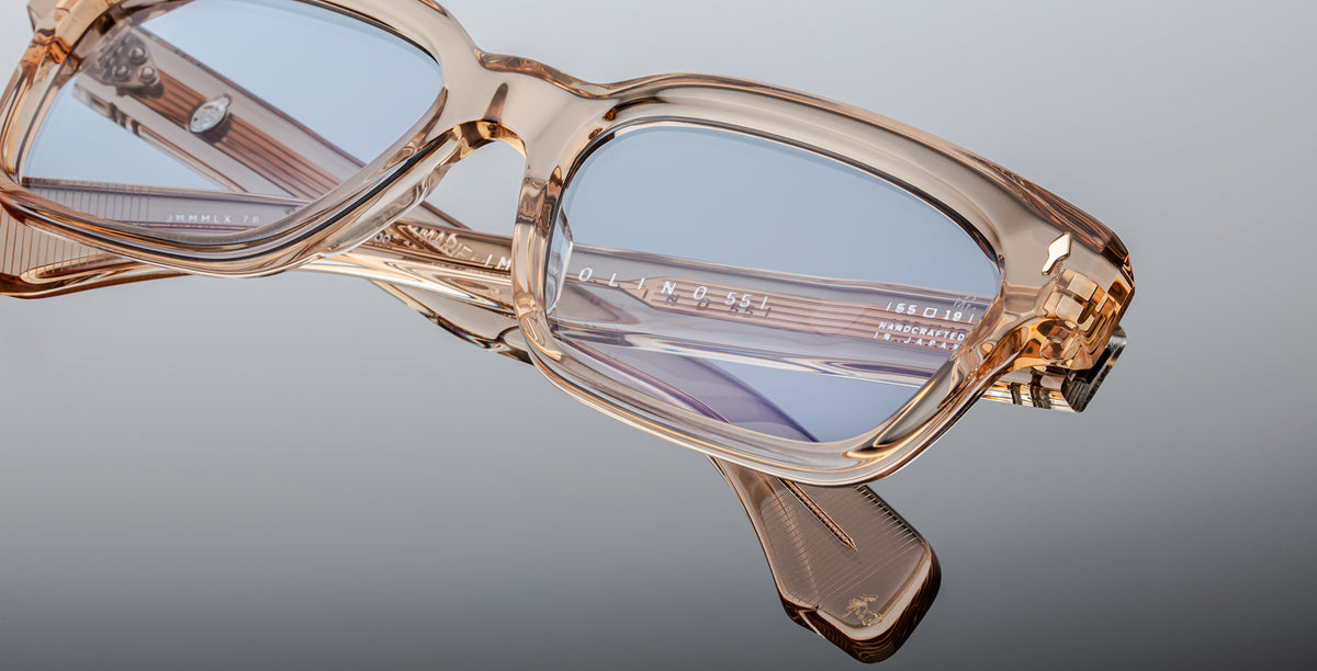 Close-up of Jacques Marie Mage Molino 55 eyeglasses in transparent beige with blue-tinted lenses, featuring detailed frame design and branding on the temple arm, set against a gray background.