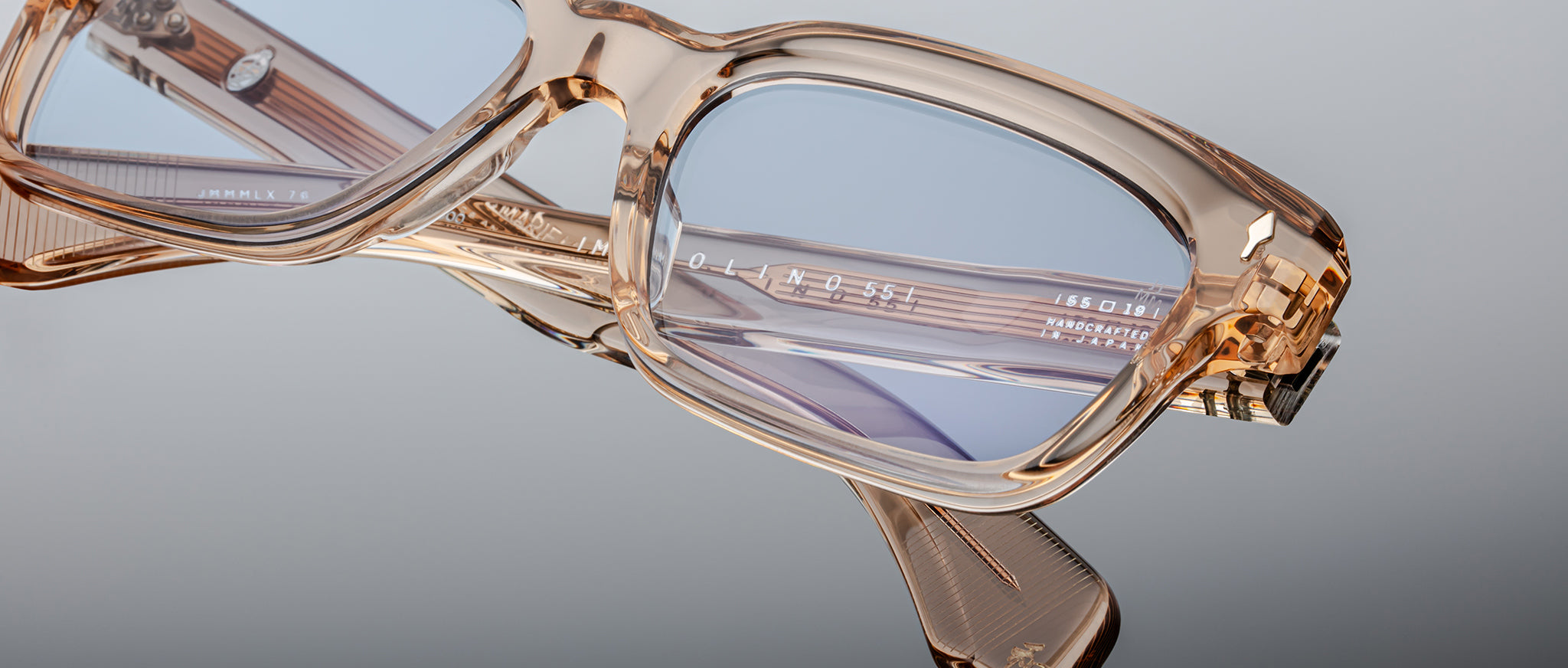 Close-up of Jacques Marie Mage Molino 55 eyeglasses in transparent beige with blue-tinted lenses, featuring detailed frame design and branding on the temple arm, set against a gray background.
