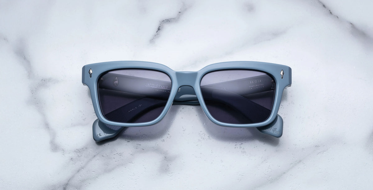 A pair of Jacques Marie Mage Molino 55 blue rectangular sunglasses with dark lenses rests on a white marble surface.