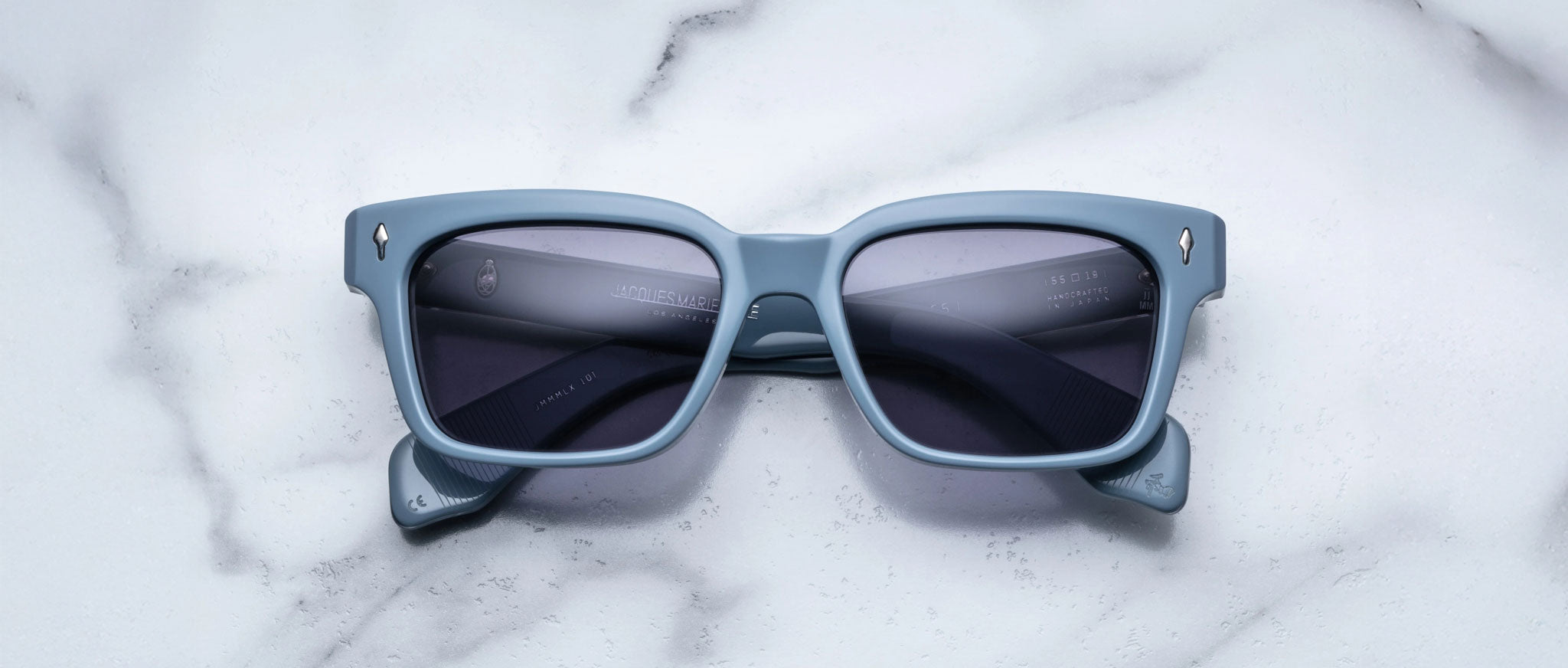 A pair of Jacques Marie Mage Molino 55 blue rectangular sunglasses with dark lenses rests on a white marble surface.