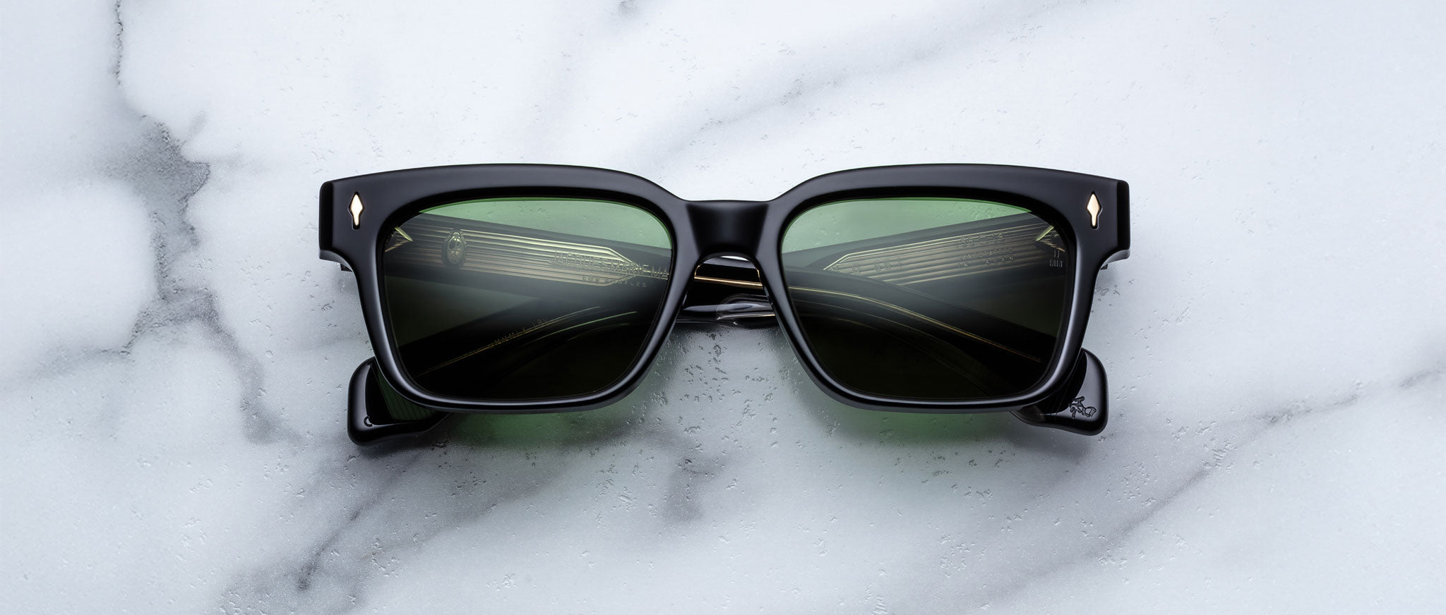 The Jacques Marie Mage Molino 55, black square-framed sunglasses with green-tinted lenses, is displayed on a white marble surface. The lenses reflect a metallic or architectural structure.