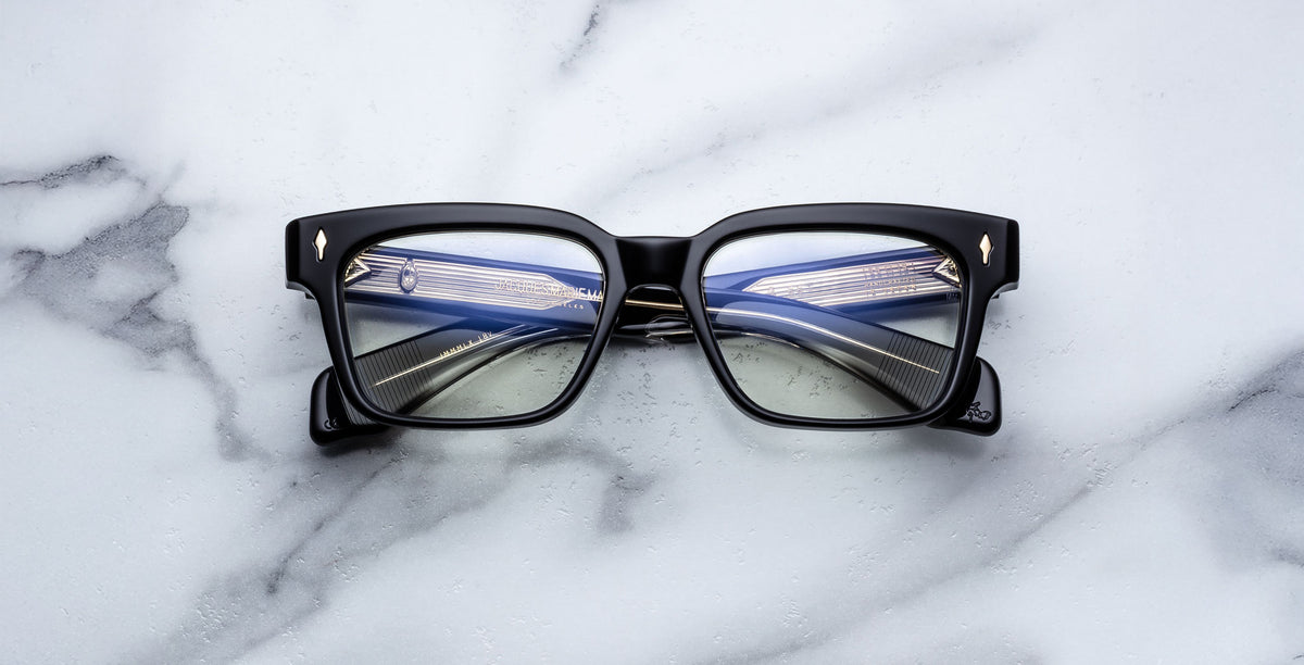 The Jacques Marie Mage Molino 55 eyeglasses, featuring black square frames with gold corner details, rest on a white marble surface. The lenses reflect the image of a modern bridge illuminated by cables and lights.