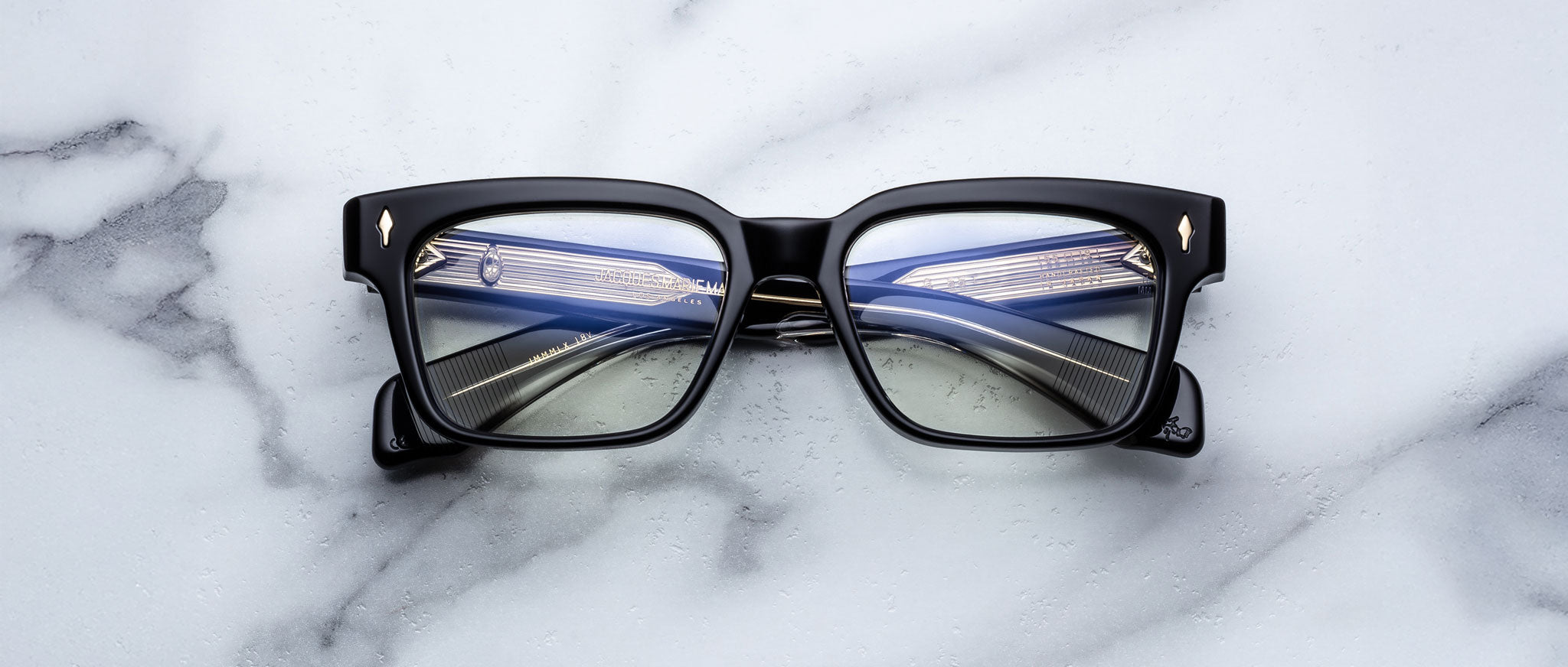 The Jacques Marie Mage Molino 55 eyeglasses, featuring black square frames with gold corner details, rest on a white marble surface. The lenses reflect the image of a modern bridge illuminated by cables and lights.