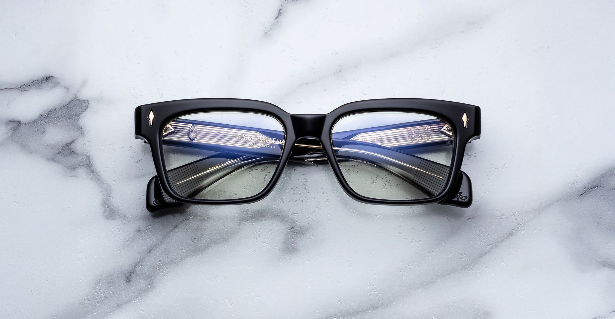 The Jacques Marie Mage Molino 55 eyeglasses, featuring black square frames with gold corner details, rest on a white marble surface. The lenses reflect the image of a modern bridge illuminated by cables and lights.