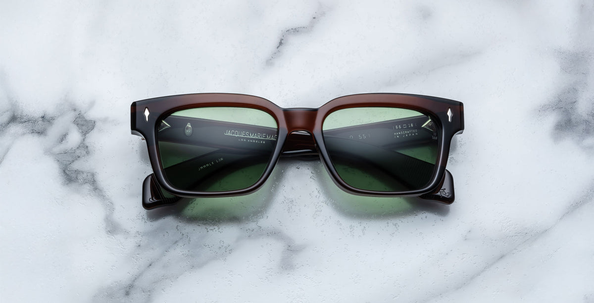 A pair of Jacques Marie Mage Molino 55 square, dark brown sunglasses with green-tinted lenses rests on a white marble surface.