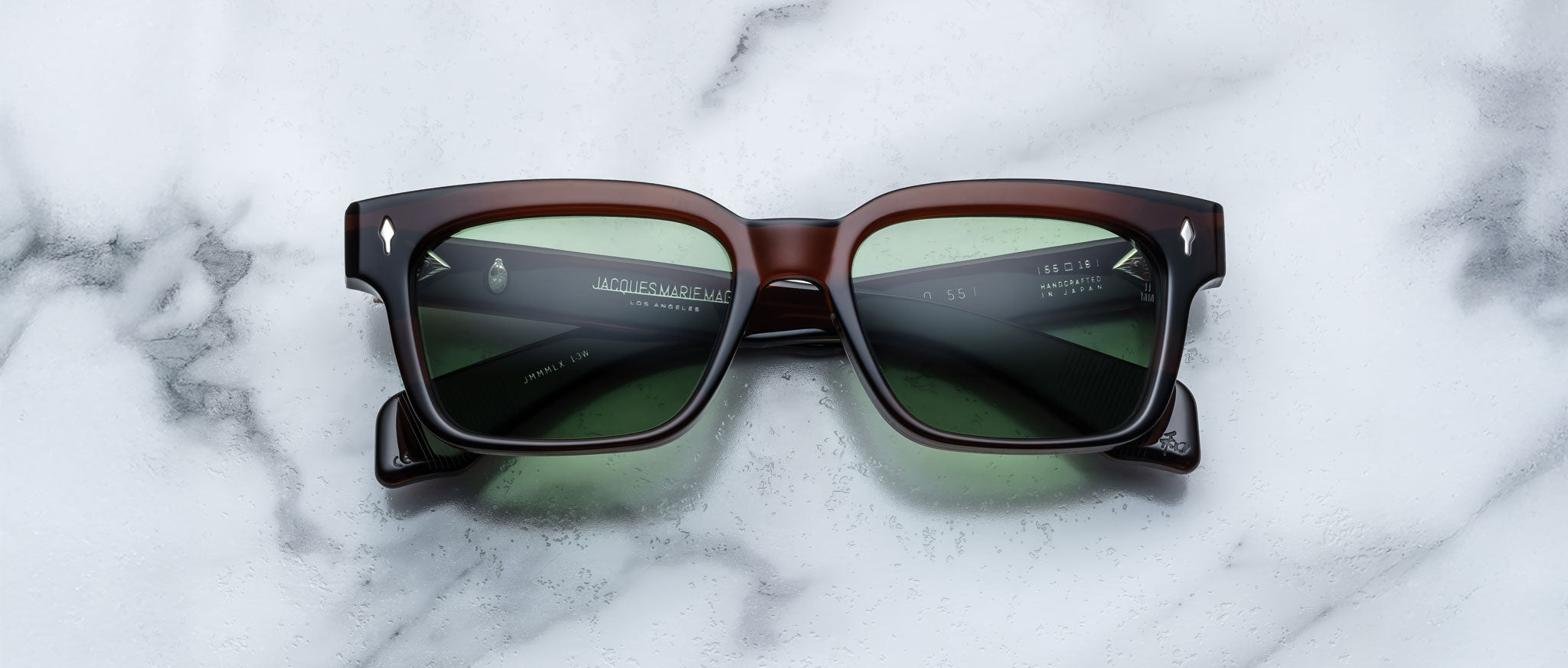 A pair of Jacques Marie Mage Molino 55 square, dark brown sunglasses with green-tinted lenses rests on a white marble surface.