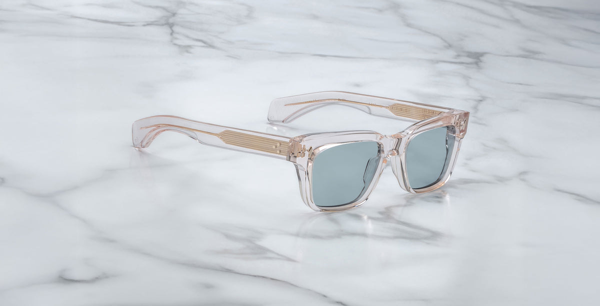 The Jacques Marie Mage Molino sunglasses in Cameo feature a clear, square-shaped frame with gold accents and blue-tinted lenses, displayed on a white marble surface.