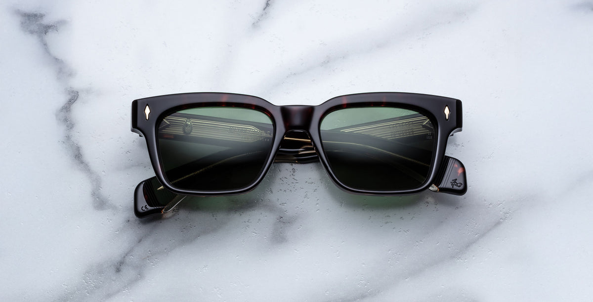 A pair of Jacques Marie Mage Molino black rectangular sunglasses with green-tinted lenses lies on a white marble surface, the lenses reflecting part of a building with windows and railings.