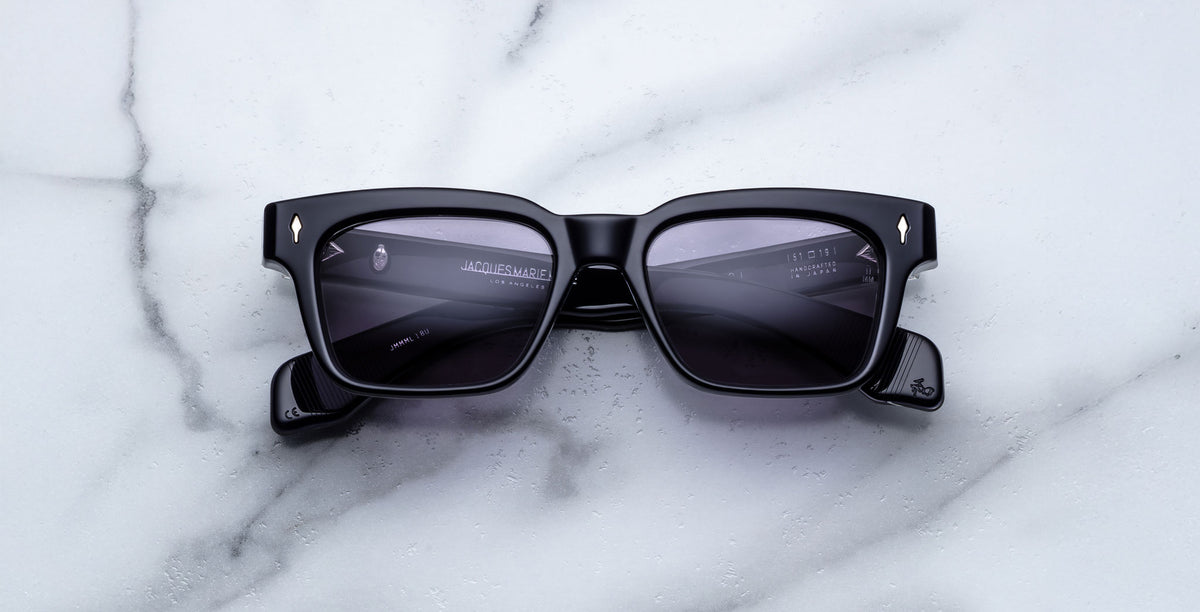 A pair of Jacques Marie Mage Molino black rectangular sunglasses with dark lenses rests on white marble, featuring silver corner accents and subtle branding on the temples.