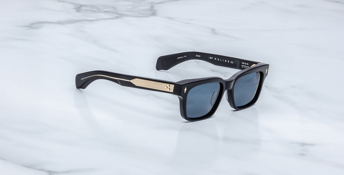 The Jacques Marie Mage Molino sunglasses feature black frames, dark lenses, and gold metallic temple accents, displayed on a white marble surface.