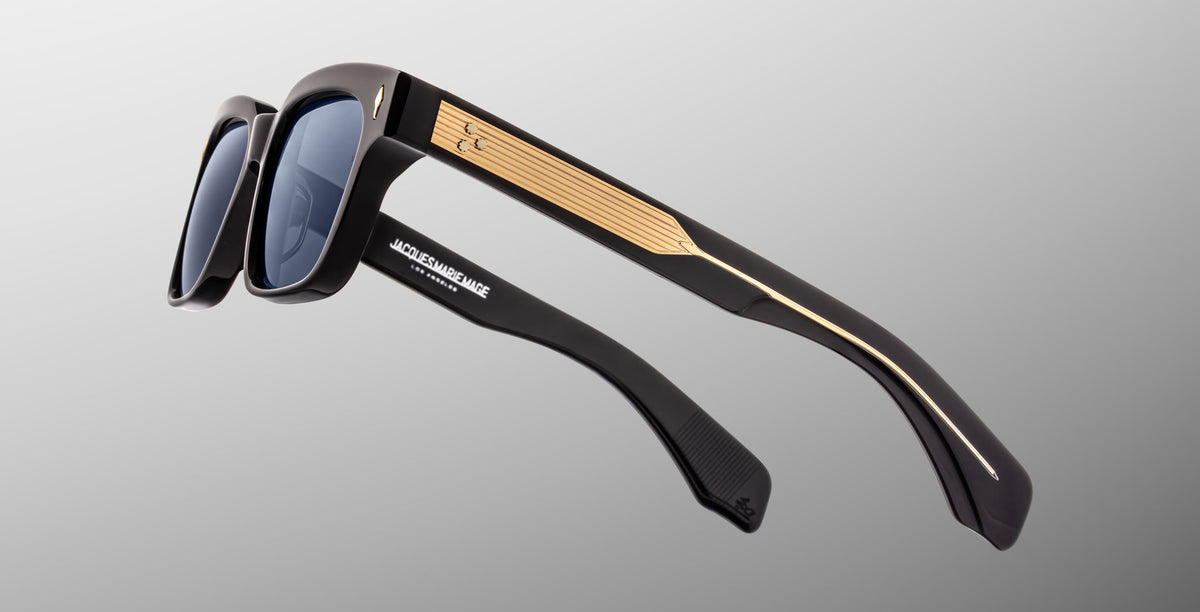 The Jacques Marie Mage Molino in Lacquer features dark blue sunglasses with dark lenses, thick arms, gold detailing on the sides, and a metallic accent near the hinges, displayed against a gradient gray background.