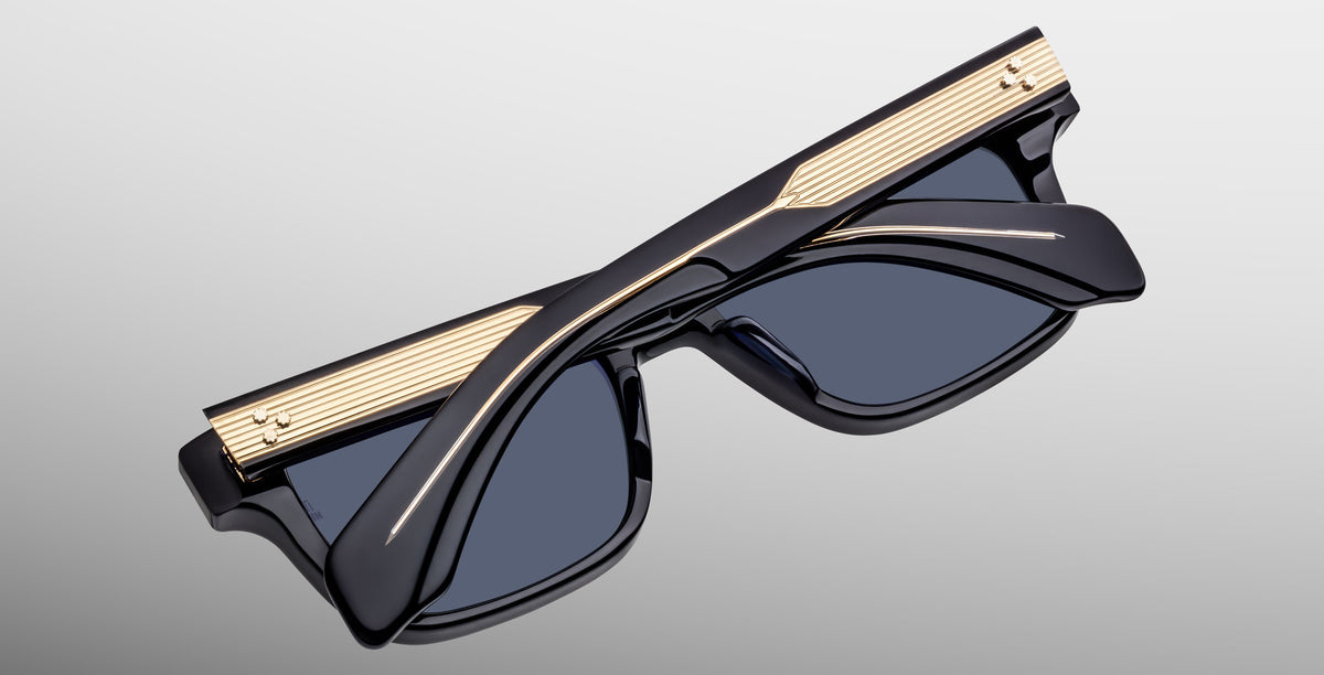 The Jacques Marie Mage Molino sunglasses in Lacquer feature dark blue frames with dark lenses, gold accents, and a ribbed top edge, shown against a light gray background.
