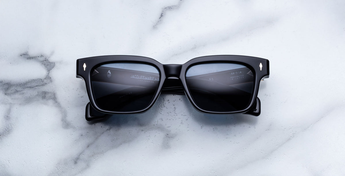 The Jacques Marie Mage Molino a pair of black square sunglasses with dark lenses, rests on a white marble surface, reflecting light and faint text in the lenses.
