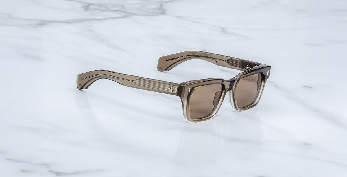 The Jacques Marie Mage Molino sunglasses feature square, brown-tinted lenses and thick, translucent brown frames, shown resting on a white marble surface.