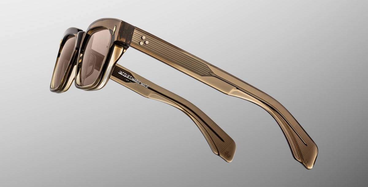 A close-up of Jacques Marie Mage Molino sunglasses in Lescot, featuring translucent brown frames, thick textured arms, and light brown tinted lenses, set against a neutral gray gradient background.