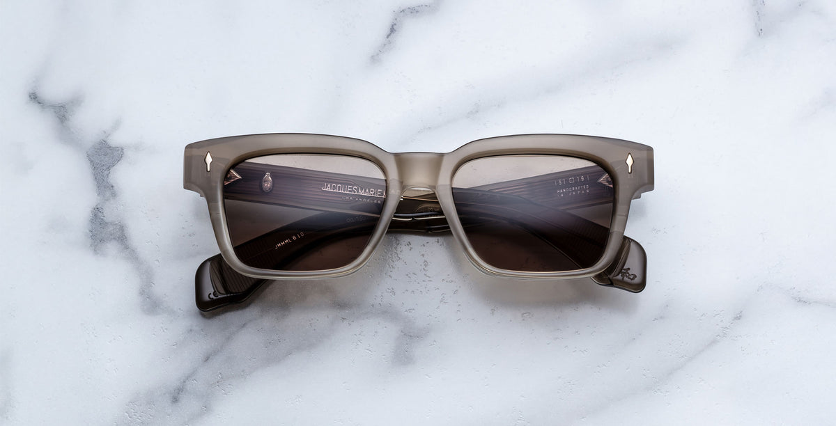 Jacques Marie Mage Molino sunglasses featuring square gray frames and dark tinted lenses, rest on a white marble surface with their arms crossed behind the lenses.