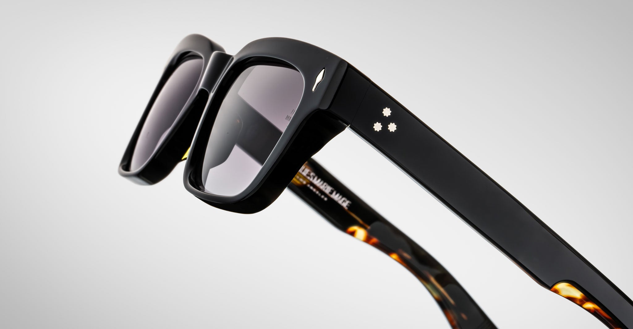 Close-up of Jacques Marie Mage Molino black sunglasses, with thick frames, two white stars on the temple, and tortoiseshell inner arms, set against a light gray background.